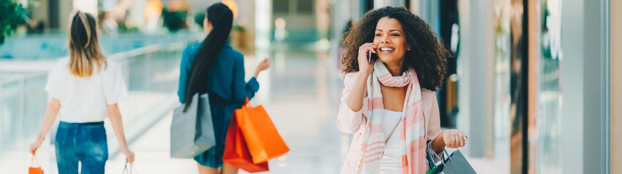 Femme souriante marchant dans un centre commercial lumineux, tenant plusieurs sacs de shopping et parlant au téléphone. En arrière-plan, d'autres clientes portent également des sacs de différentes couleurs. L’image illustre une expérience de shopping dynamique et connectée.