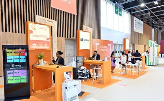 Startup Village exhibition stand with several companies on display, work tables, orange panels and an area dedicated to professional discussions.