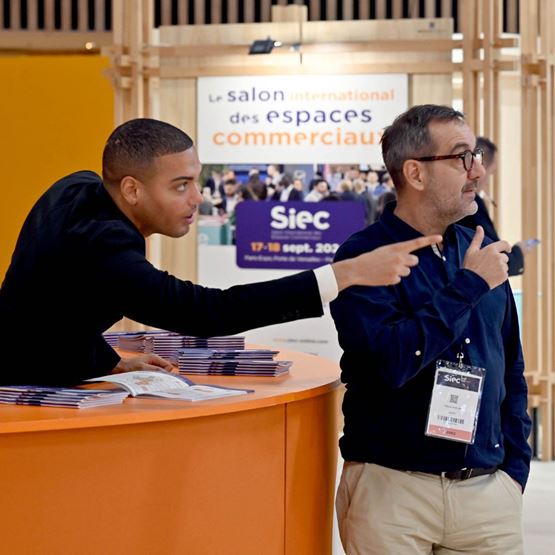 A Siec trade show staff member assists a visitor by pointing in a direction, standing at an orange information desk with a show banner in the background.